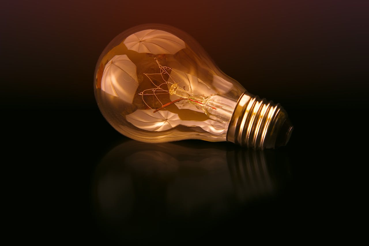 gallery-4 A detailed close-up of a light bulb showcasing its intricate filament and glowing glass against a dark reflective surface.