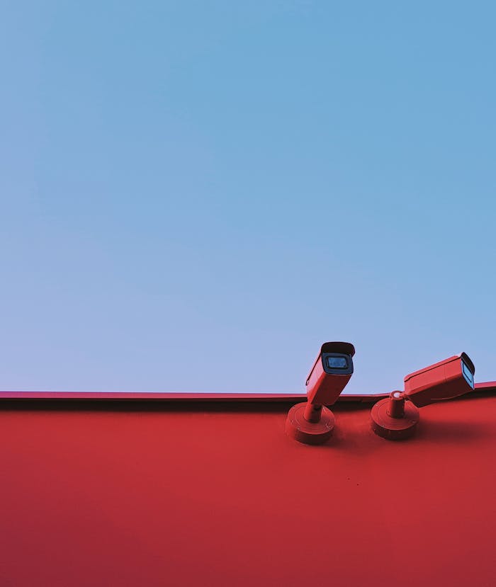 ours-journey Low-angle shot of red CCTV cameras on a red wall with a clear blue sky background.